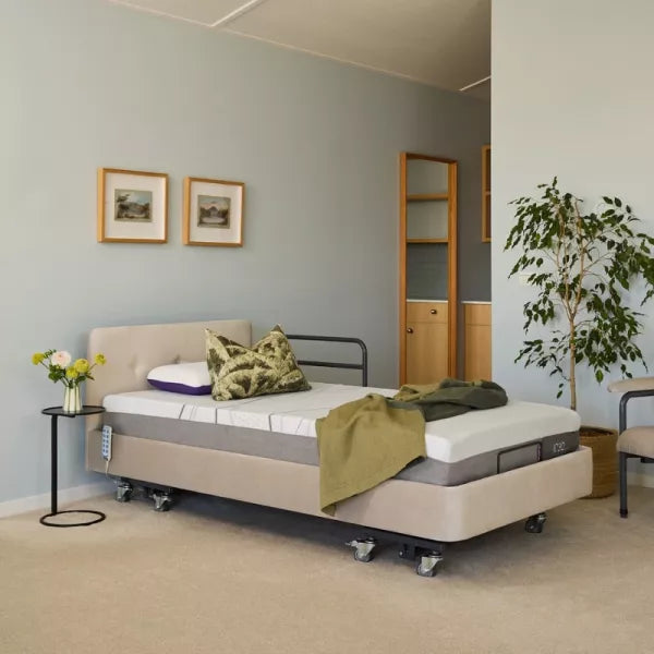 An adjustable electric hospital bed with a beige upholstered base and side rails, shown in a styled bedroom setting with a side table, framed pictures, a plant, and a chair.